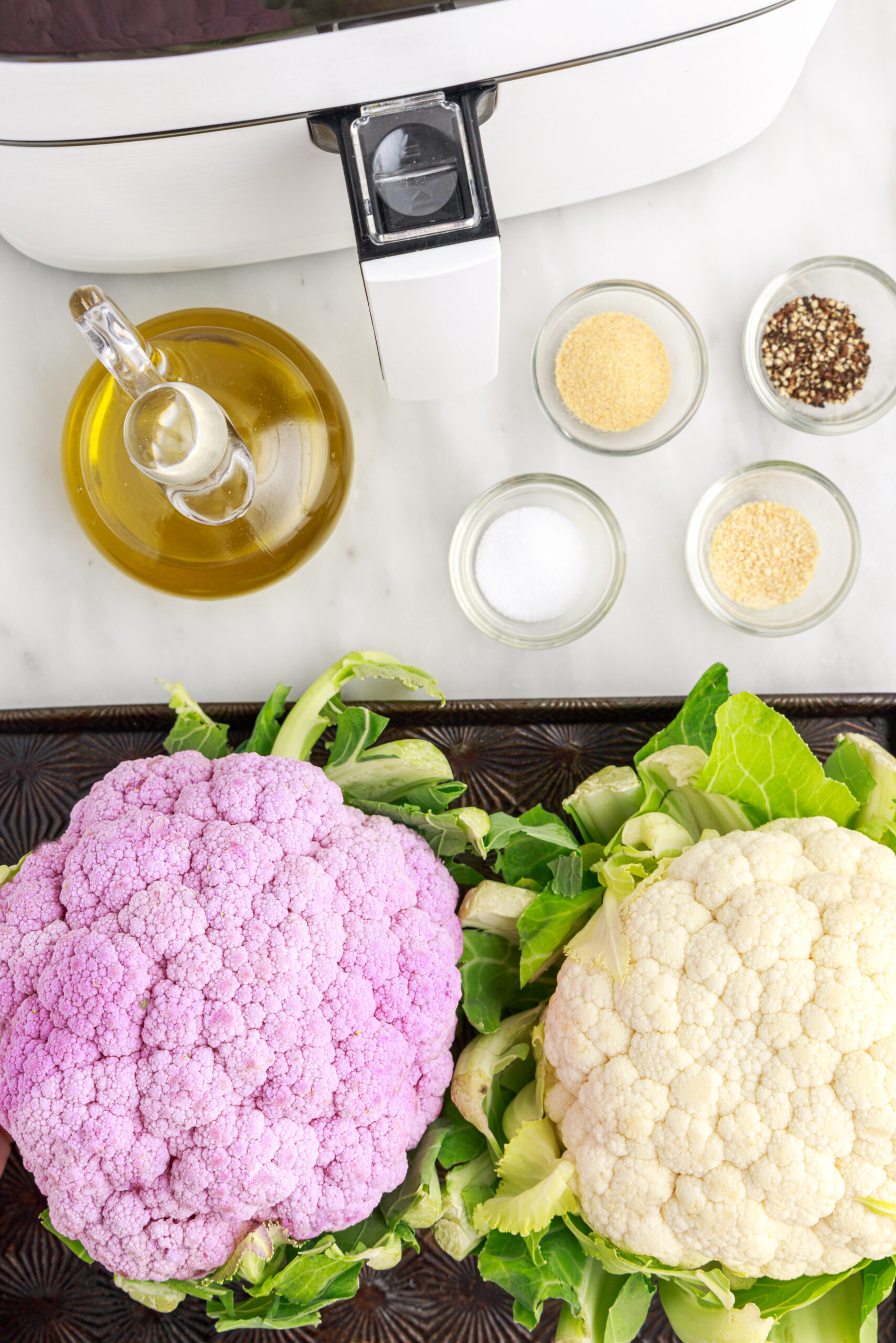 A head of purple cauliflower, a head of white cauliflower, and bowls of garlic powder, onion powder, salt, pepper, and a jar of olive oil