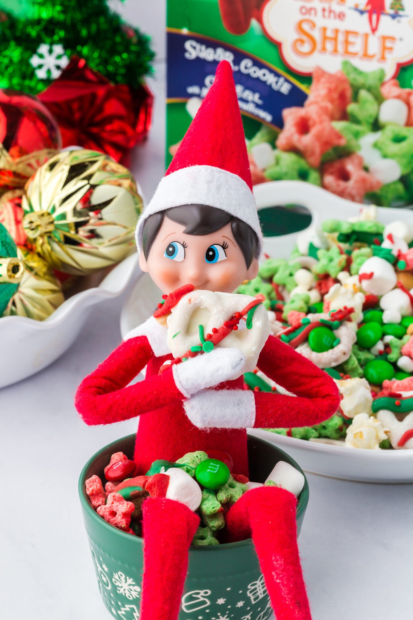Elf on the Shelf doll sitting on a cup of festive Christmas snack mix