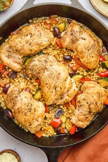 One pan Mediterranean chicken and couscous in a skillet with juicy chicken thighs, pearl couscous, vegetables, olives, and fresh herbs