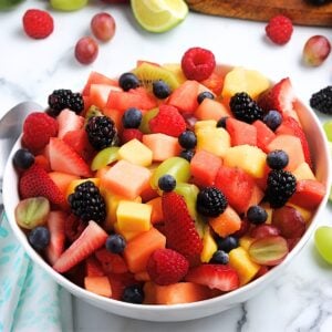 Best summer fruit salad in a white bowl filled with watermelon, cantaloupe, strawberries, grapes, blueberries, raspberries, blackberries, mango, and kiwi chunks on a marble background