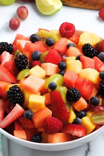 Best summer fruit salad in a white bowl filled with watermelon, cantaloupe, strawberries, grapes, blueberries, raspberries, blackberries, mango, and kiwi chunks on a marble background